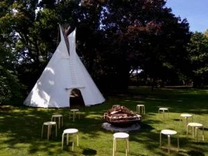 Een sfeervolle witte tipi tent op een groen grasveld, compleet met vuurschaal en houten zitplaatsen voor een verjaardagsfeest aan de bosrand in Gelderland